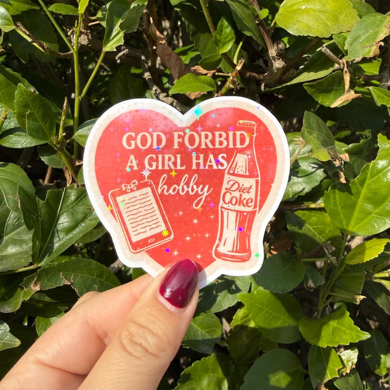 "God forbid a girl has a hobby" in pink and white. The sticker features illustrations of a Diet Coke bottle and an e-reader.
