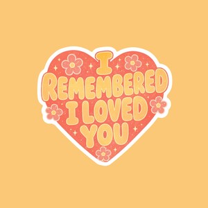 May include: A heart-shaped sticker with a coral-red background featuring the yellow text I REMEMBERED I LOVED YOU. The design includes small yellow flowers and tiny star accents, all outlined in a thick white border against a solid yellow background.