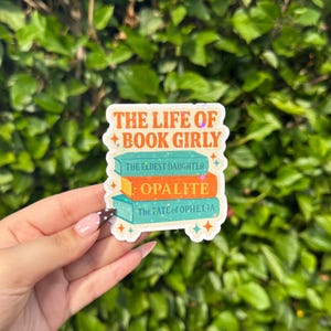 May include: A sticker with the text "THE LIFE OF BOOK GIRLY" and three stacked books. The books are teal and orange, with titles including "THE ELDEST DAUGHTER" and "OPALITE". The sticker has a white border and star accents.