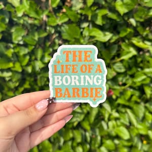 May include: A sticker with the text "THE LIFE OF A BORING BARBIE" in orange and white on a light green background. The sticker is held in front of a green leafy background.