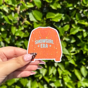 May include: An orange sticker featuring a cartoon illustration of a cardigan sweater. The text "SHOWGIRL ERA" is printed on the sweater in white and teal. The sticker has a glittery effect with star accents. The background is a lush green foliage.