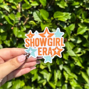 May include: A white sticker with the words "SHOWGIRL ERA" in orange text. The sticker has orange and light blue star accents. The sticker is held in a hand with a polka dot nail design.