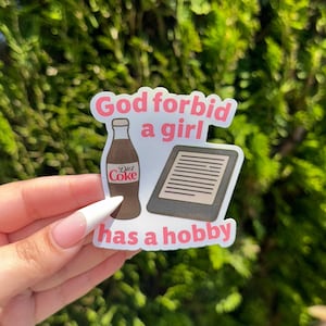 May include: A sticker with the text "God forbid a girl has a hobby" in pink and white. The sticker features illustrations of a Diet Coke bottle and an e-reader.