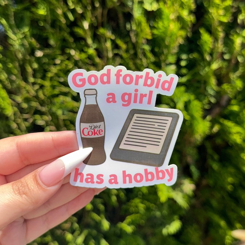 May include: A sticker with the text "God forbid a girl has a hobby" in pink and white. The sticker features illustrations of a Diet Coke bottle and an e-reader.