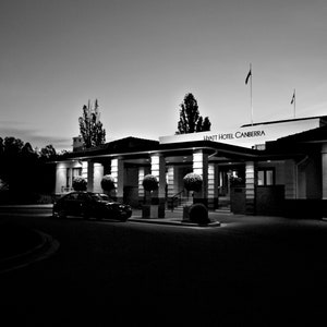 以下が含まれることがあります： 夕暮れ時のハイアットホテルキャンベラの白黒写真。ホテルの入り口はライトアップされ、前に車が停まっています。ホテルの名前は入り口の上に表示されています。