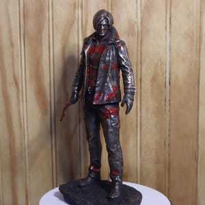 Pode incluir: Estatueta detalhada de uma pessoa com jaqueta e calças de couro, salpicada com tinta vermelha. A figura está segurando uma lâmina e está em uma base escura e texturizada. A estátua tem um acabamento metálico.
