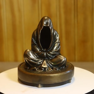 May include: A bronze-toned figurine of a hooded, faceless figure sitting in a cross-legged position. The sculpture features draped fabric details and rests upon a circular, elevated base with a smooth, metallic finish and dark patina accents.
