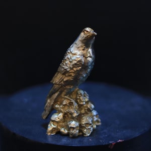 Puede incluir: Figura dorada de un ave posada sobre una base de calaveras. El ave tiene texturas detalladas de plumas y una mirada enfocada. La escultura está sobre un fondo oscuro, resaltando los intrincados detalles de la figura aviar.