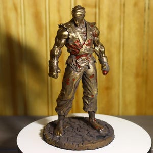Op de afbeelding: Een bronzen beeldje van een gespierde figuur in vechthouding, met een blinddoek en bloedrode accenten. Het beeldje staat op een cirkelvormige basis met een getextureerd oppervlak.