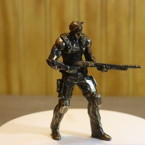 May include: A metallic silver-toned figurine of a soldier in tactical gear, standing in a combat stance while holding a long rifle. The figure features detailed armor plating on the chest, shoulders, and legs, with a focused posture.