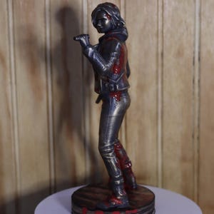 May include: A metallic figurine of a person in a jacket and pants, with red accents suggesting blood. The figure is standing on a round, wooden-textured base. The overall aesthetic is a blend of action and artistry.
