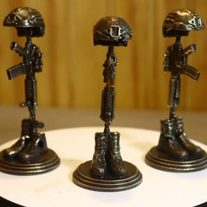 May include: Three bronze-colored metal sculptures of a fallen soldier memorial, each featuring a pair of combat boots at the base, a rifle standing vertically in the center, and a tactical helmet resting on top, all mounted on a circular base.