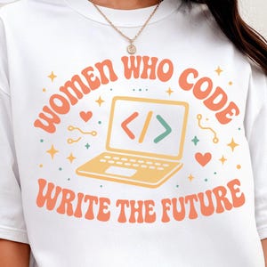 May include: White t-shirt with the text "WOMEN WHO CODE WRITE THE FUTURE" in orange. The design includes a yellow laptop with code symbols, hearts, and stars. The top left corner has a white rectangle with the words "WHITE COMFORT COLORS".