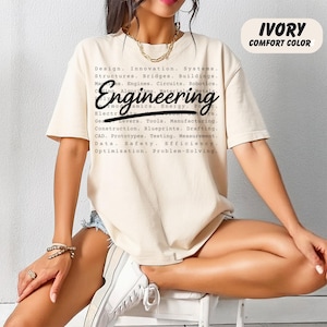 May include: Ivory-colored t-shirt with the word "Engineering" in a bold, stylized font. The shirt features a list of engineering-related terms. The text "IVORY COMFORT COLOR" is printed at the top right.