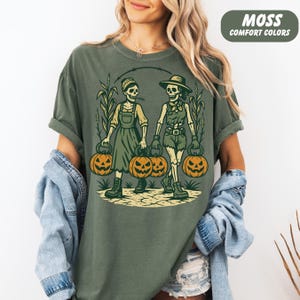 front-Moss Comfort Colors T-shirt featuring two vintage-style skeleton women dressed in farm outfits, holding jack-o'-lanterns with corn stalks in the background. The design has a rustic fall and Halloween aesthetic.
