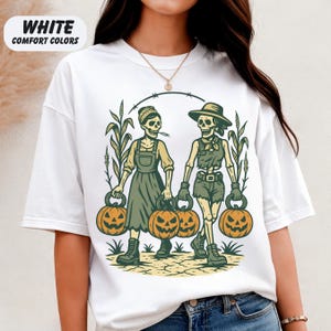 front-White Comfort Colors T-shirt featuring two vintage-style skeleton women dressed in farm outfits, holding jack-o'-lanterns with corn stalks in the background. The design has a rustic fall and Halloween aesthetic.