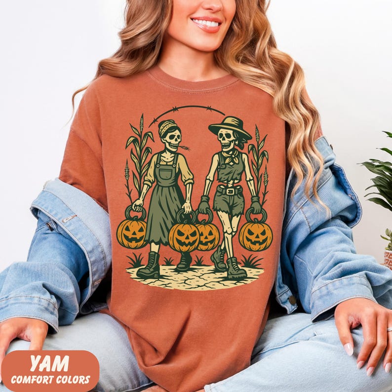 front-Yam Comfort Colors T-shirt featuring two vintage-style skeleton women dressed in farm outfits, holding jack-o'-lanterns with corn stalks in the background. The design has a rustic fall and Halloween aesthetic.