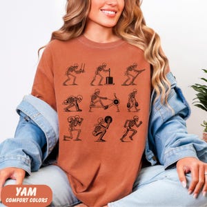 May include: Rust-colored t-shirt featuring a black graphic of skeletons exercising with weights, rowing machines, and other gym equipment. The text "YAM COMFORT COLORS" is at the bottom. The shirt is a comfortable, casual style.