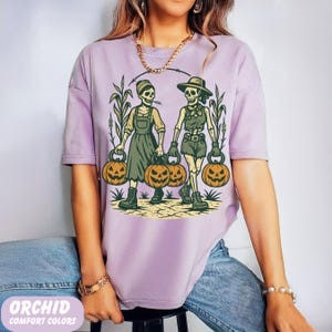 front-Orchid Comfort Colors T-shirt featuring two vintage-style skeleton women dressed in farm outfits, holding jack-o'-lanterns with corn stalks in the background. The design has a rustic fall and Halloween aesthetic.