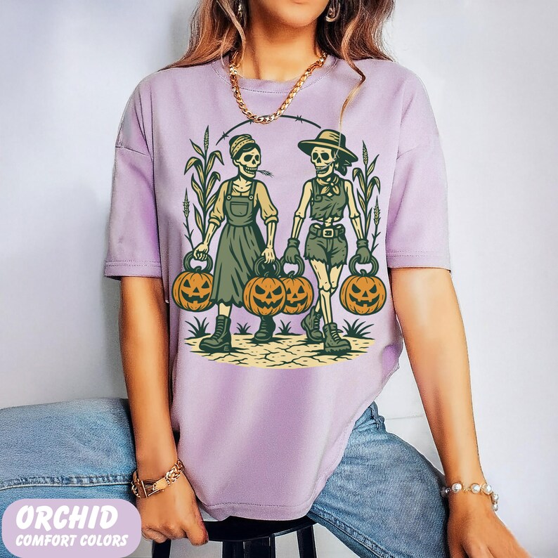 front-Orchid Comfort Colors T-shirt featuring two vintage-style skeleton women dressed in farm outfits, holding jack-o'-lanterns with corn stalks in the background. The design has a rustic fall and Halloween aesthetic.