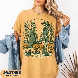 front-Mustard Comfort Colors T-shirt featuring two vintage-style skeleton women dressed in farm outfits, holding jack-o'-lanterns with corn stalks in the background. The design has a rustic fall and Halloween aesthetic.