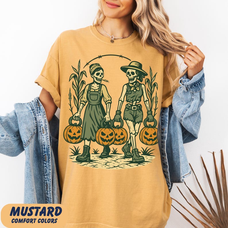 front-Mustard Comfort Colors T-shirt featuring two vintage-style skeleton women dressed in farm outfits, holding jack-o'-lanterns with corn stalks in the background. The design has a rustic fall and Halloween aesthetic.