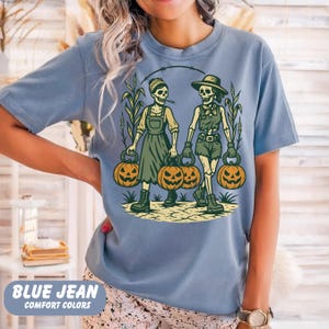 front-Blue Jean Comfort Colors T-shirt featuring two vintage-style skeleton women dressed in farm outfits, holding jack-o'-lanterns with corn stalks in the background. The design has a rustic fall and Halloween aesthetic.