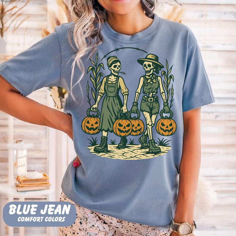 front-Blue Jean Comfort Colors T-shirt featuring two vintage-style skeleton women dressed in farm outfits, holding jack-o'-lanterns with corn stalks in the background. The design has a rustic fall and Halloween aesthetic.
