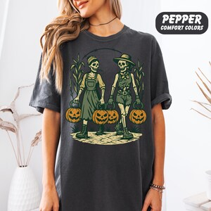front-Pepper Comfort Colors T-shirt featuring two vintage-style skeleton women dressed in farm outfits, holding jack-o'-lanterns with corn stalks in the background. The design has a rustic fall and Halloween aesthetic.
