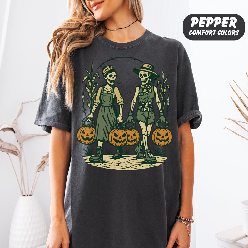 front-Pepper Comfort Colors T-shirt featuring two vintage-style skeleton women dressed in farm outfits, holding jack-o'-lanterns with corn stalks in the background. The design has a rustic fall and Halloween aesthetic.