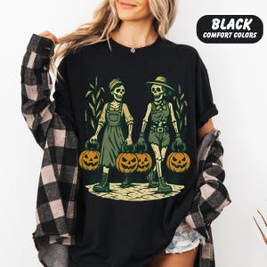 front-Black Comfort Colors T-shirt featuring two vintage-style skeleton women dressed in farm outfits, holding jack-o'-lanterns with corn stalks in the background. The design has a rustic fall and Halloween aesthetic.