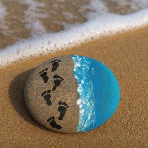 May include: A pebble painted with a beach scene. One side has black footprints, the other is painted blue to represent the sea. White paint is used to depict waves. The pebble rests on sand.