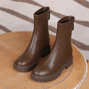 Puede incluir: Un par de botines marrones con punta redondeada y suela gruesa. Las botas tienen una cremallera en la parte trasera y están hechas de un material liso. Las botas están sobre una mesa de madera.