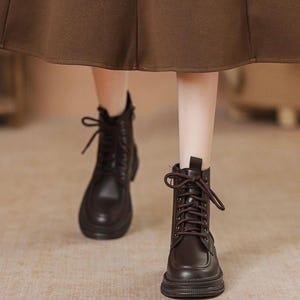 Könnte beinhalten: Dunkelbraune Leder-Schnürstiefel mit runder Spitze und dicker Sohle. Die Stiefel haben ein klassisches Design mit mehreren Ösen für die Schnürsenkel. Das Bild zeigt die Stiefel von den Knöcheln abwärts, mit einem braunen Rock im Hintergrund.