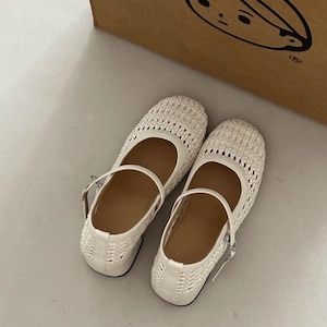 May include: A pair of white woven Mary Jane flats with a strap and buckle closure. The shoes have a rounded toe and a small heel. The woven design allows for ventilation. The shoes are sitting on a light-colored surface.