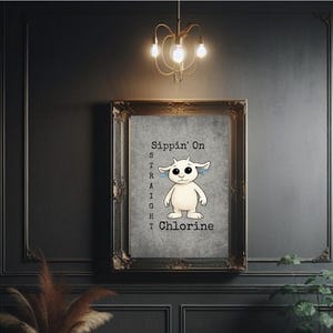 May include: Framed artwork featuring a cartoon creature with large eyes and blue accents. The text "Sippin' On Straight Chlorine" is vertically aligned on the left side of the image. The artwork is displayed in a dark, ornate frame against a dark wall.