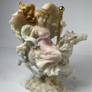 May include: A porcelain figurine of an angel riding a white carousel horse. The angel wears a pink dress and has flowing blonde hair and feathered wings. The horse is adorned with pink roses, and the base features a cloud-like design with more roses.