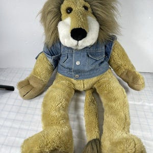 May include: A plush lion toy with tan fur, a long brown mane, and a white muzzle featuring a black nose. The lion is wearing a blue denim vest with two silver-colored buttons on the front. The toy has a long tail with a tuft of brown fur at the end.