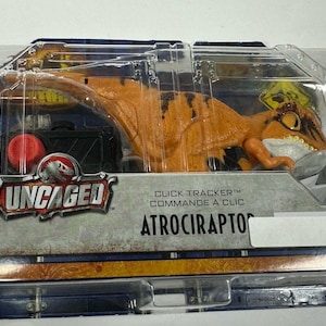 May include: An orange and brown Atrociraptor dinosaur toy in a clear plastic package. The package includes the text "UNCAGED" and "ATROCIRAPTOR". A red button and a black box are also visible.