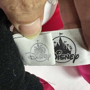 Op de afbeelding: Close-up van een label met het Disney-logo en tekst. Het label is bevestigd aan een zwart en roze pluche speelgoed. Het Disney-logo toont een kasteelsilhouet. Het label is wit met grijze tekst en een grijs logo.
