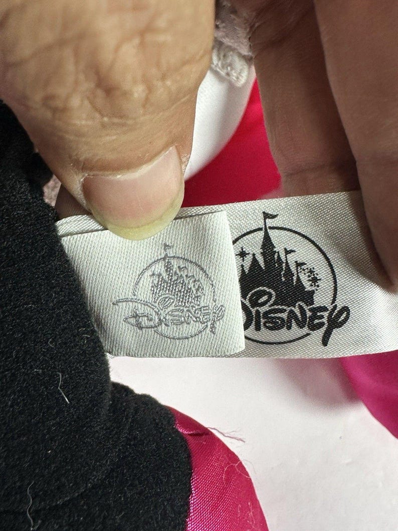 Op de afbeelding: Close-up van een label met het Disney-logo en tekst. Het label is bevestigd aan een zwart en roze pluche speelgoed. Het Disney-logo toont een kasteelsilhouet. Het label is wit met grijze tekst en een grijs logo.