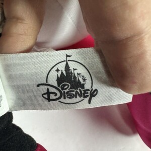 Op de afbeelding: Een close-up van een Disney-label. Het label is lichtgrijs met het Disney-logo in zwart. Het logo toont een kasteelsilhouet met het woord "Disney" eronder. Het label wordt door een hand vastgehouden.