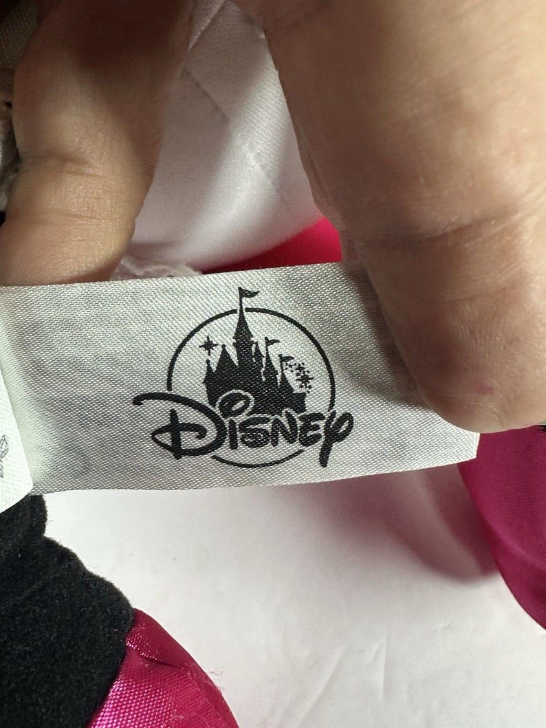 Op de afbeelding: Een close-up van een Disney-label. Het label is lichtgrijs met het Disney-logo in zwart. Het logo toont een kasteelsilhouet met het woord "Disney" eronder. Het label wordt door een hand vastgehouden.