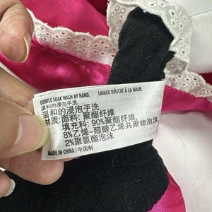 Op de afbeelding: Close-up van een pluche speelgoed met zwart en wit stoffen detail en roze satijnen stof. Een label met Engelse en Chinese tekst luidt "GENTLE SOAK WASH BY HAND" en "MADE IN CHINA".