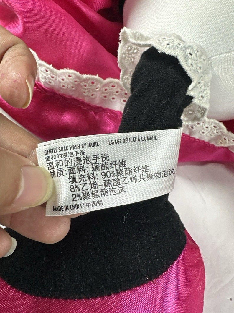 Op de afbeelding: Close-up van een pluche speelgoed met zwart en wit stoffen detail en roze satijnen stof. Een label met Engelse en Chinese tekst luidt "GENTLE SOAK WASH BY HAND" en "MADE IN CHINA".