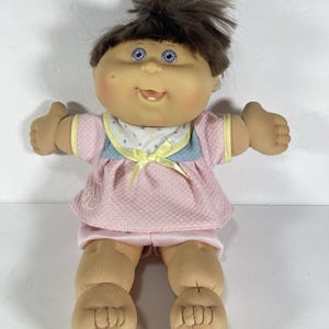 May include: A soft-bodied doll with brown hair, blue eyes, and a dimpled face. The doll wears a pink textured top with a blue yoke, a white collar with colorful polka dots, a yellow ribbon bow, and pink shorts. A small heart logo is on the top.
