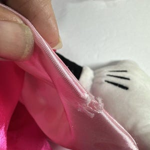 Op de afbeelding: Close-up van een roze satijnen strik met een kleine scheur, onderdeel van een pluchen speelgoed. De strik wordt vastgehouden door een hand, waardoor de textuur en kleur van de stof zichtbaar worden. Een witte handschoen met zwarte lijnen is zichtbaar.