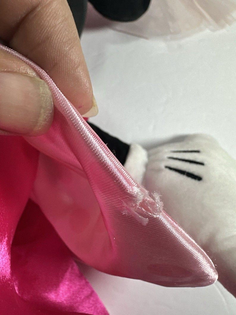 Op de afbeelding: Close-up van een roze satijnen strik met een kleine scheur, onderdeel van een pluchen speelgoed. De strik wordt vastgehouden door een hand, waardoor de textuur en kleur van de stof zichtbaar worden. Een witte handschoen met zwarte lijnen is zichtbaar.
