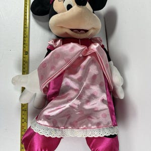 Op de afbeelding: Een pluche Minnie Mouse-pop, voornamelijk roze en zwart, met een roze jurk en strik. De pop heeft witte handschoenen en een glimlachende uitdrukking. De pop is ongeveer 61 cm hoog.