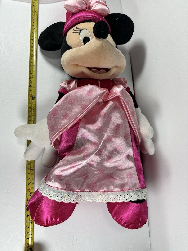 Op de afbeelding: Een pluche Minnie Mouse-pop, voornamelijk roze en zwart, met een roze jurk en strik. De pop heeft witte handschoenen en een glimlachende uitdrukking. De pop is ongeveer 61 cm hoog.